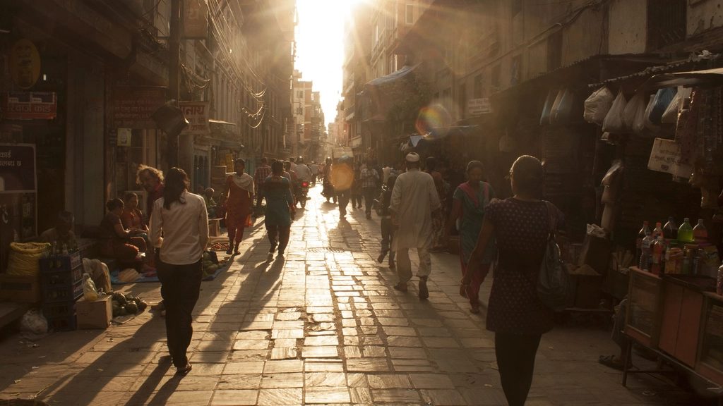 A narrow Indian market lane with shoppers backlit by low sun
