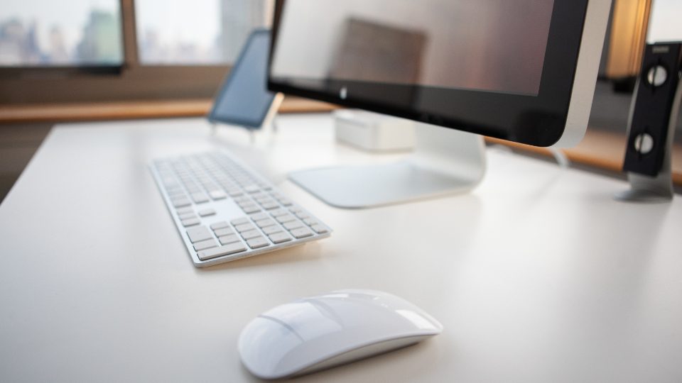 A quiet home office desk with a monitor, keyboard and phone, lit by daylight